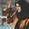 photo of a person in a headscarf standing beside a chainlink fence looking upset