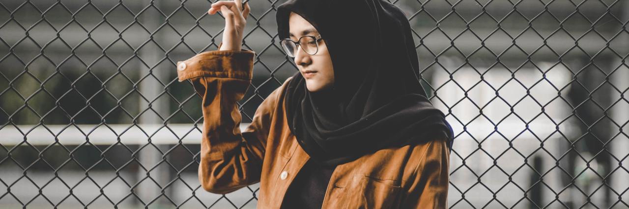 photo of a person in a headscarf standing beside a chainlink fence looking upset