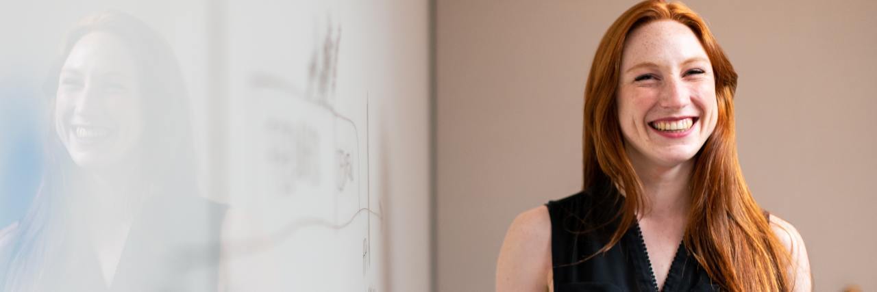 5 Job Ideas for People Living With ADHD A woman smiling next to a white board, marker in her hand. She has long red hair and is wearing a black blouse.