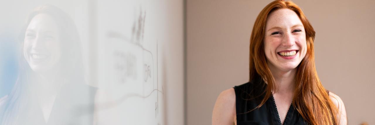 A woman smiling next to a white board, marker in her hand. She has long red hair and is wearing a black blouse.