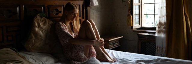 photo of a woman sitting on bed in darkness next to a windoe where the light in shining in