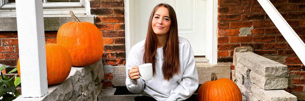 Photo of contributor sitting on front porch steps holding coffee mug, surrounded by three pumpkins