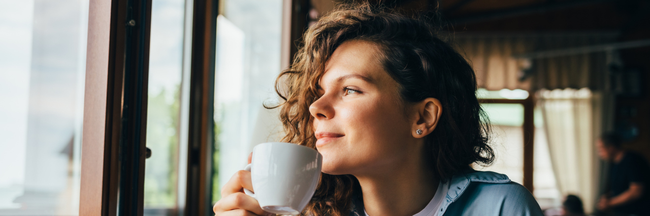 What My Mornings Are Like With Milder Depression and Anxiety Smiling calm young woman drinking coffee looking out the window sitting at table in restaurant.