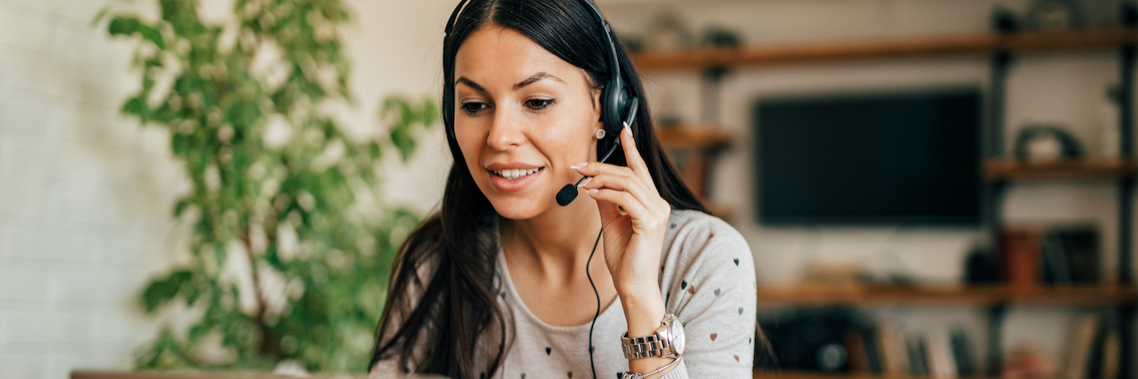 Remote Job Opportunities for People With Disabilities - September 2021 A woman sitting at her kitchen table in front of the computer with her headset on, working from home.