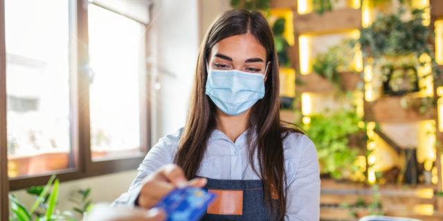 Waitress holding credit card reader machine and wearing protective face mask.