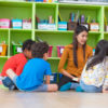 Asian female teacher teaching mixed race diversity group of kids reading book sitting on library floor in classroom