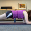A young woman with brown curly hair holds a plank on a purple matt. She wears a purple tank top and black gym shorts with a white stripe running across.