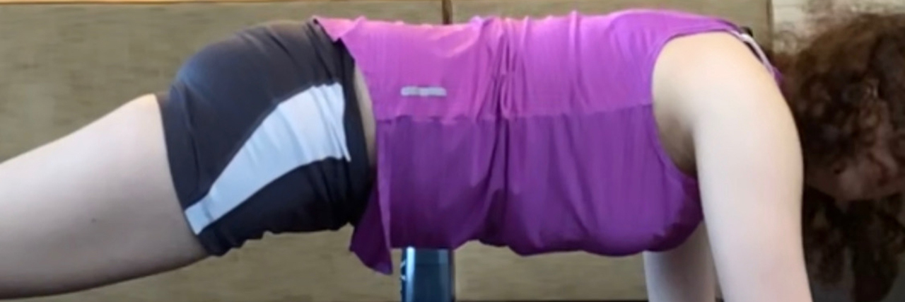 Enjoying Fitness on My Own Terms as an Autistic Adult A young woman with brown curly hair holds a plank on a purple matt. She wears a purple tank top and black gym shorts with a white stripe running across.