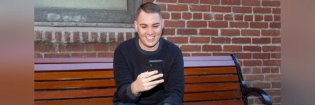 4 Tips for Coping With Job Search Anxiety Author, young man, sitting on a bench outside looking at his phone smiling