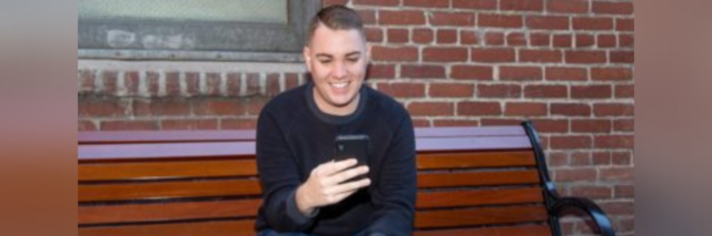 Author, young man, sitting on a bench outside looking at his phone smiling