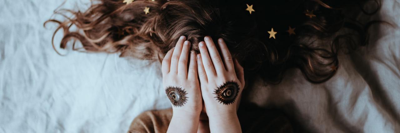 photo of young woman lying on bed covering her eyes with hands, eye tattoos on her hands