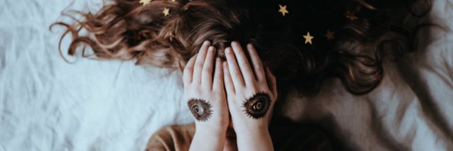 photo of young woman lying on bed covering her eyes with hands, eye tattoos on her hands