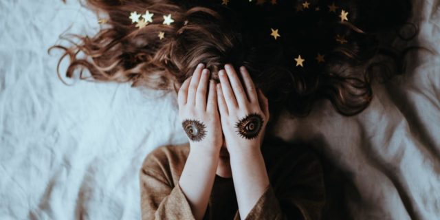 Aphantasia: How Being Unable to Visualize Anything Affects My Mental Health photo of young woman lying on bed covering her eyes with hands, eye tattoos on her hands
