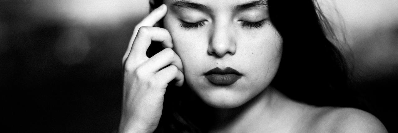 Black and white photo of a person with their eyes closed and their hand raised to their cheek, like they are trying to remember something.