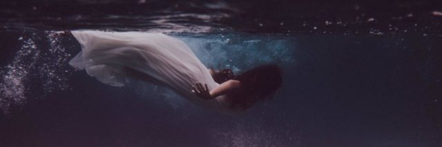 photo of a woman in a white dress swimming underwater with statues below her