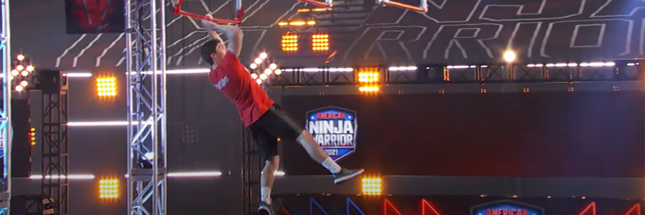 A man with a red shirt hanging at an obstacle at Ninja Warriors.