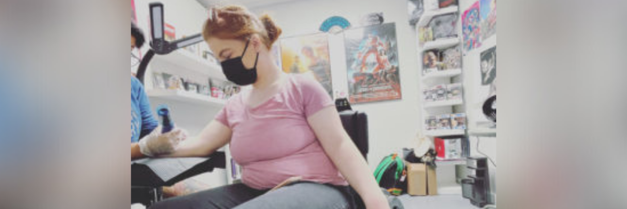 Author; young woman in a wheelchair with a mask on, arm out as a tattoo artist works