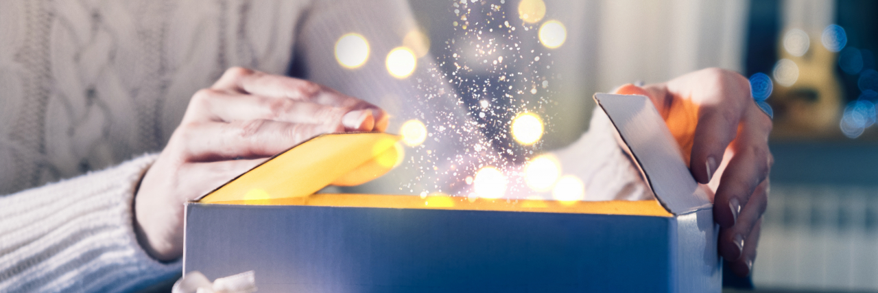 A woman in a white knit sweater opens a cardboard box surrounded by lights. There appears to be magic dust coming out of the box.