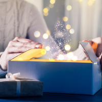 A woman in a white knit sweater opens a cardboard box surrounded by lights. There appears to be magic dust coming out of the box.