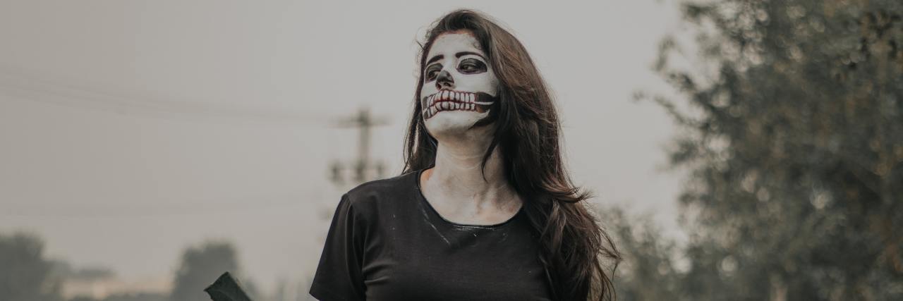 Hey Retailers, My Mental Illness Is Not Your Halloween Costume Young woman sitting outside with her arms behind her back, face painted like a skeleton
