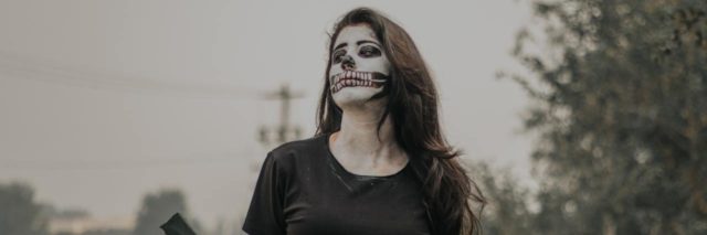 Young woman sitting outside with her arms behind her back, face painted like a skeleton