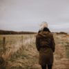 Back of a woman in a long brown jacket walking outside on a dirt road