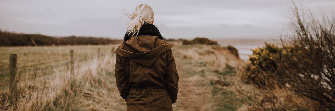 What Would Happen If I Saw My Attacker Again Back of a woman in a long brown jacket walking outside on a dirt road