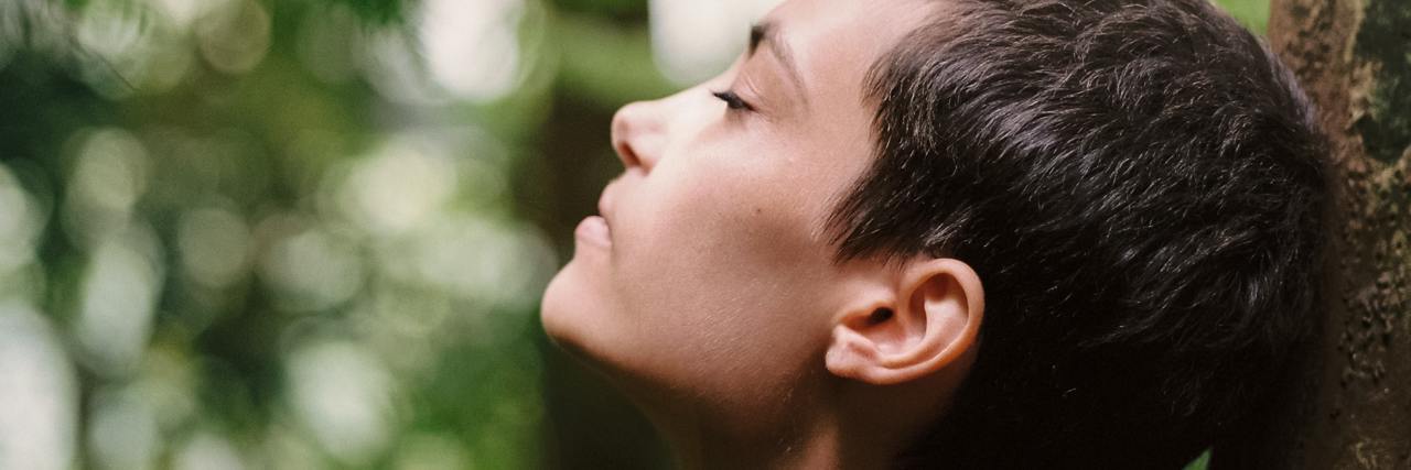 Meditation and Mindfulness May Not Be Suitable for Trauma Survivors photo of a woman with short hair leaning her head back against a tree in a forest with her eyes closed