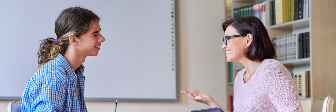Lessons I've Learned as a Social Worker for People With Disabilities A woman with short black hair wearing a blouse and slacks speaks with a young adult man with curly hair in a ponytail.