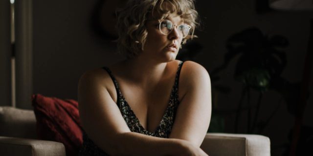 How the ‘It's Bigger Than Me’ Campaign Is Harming Fat People for Profit photo of a woman sitting on a couch in near darkness looking serious