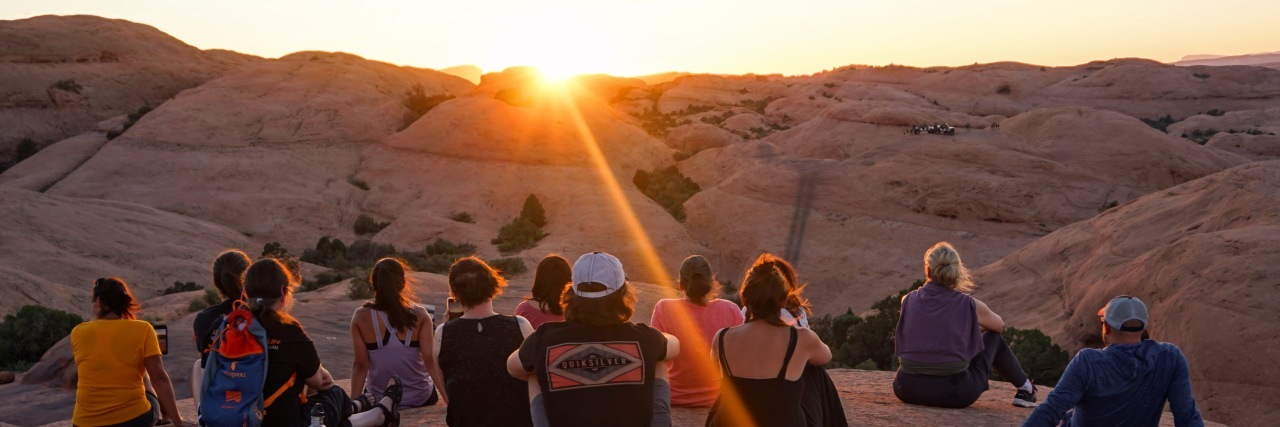 Dear 'Invisible Army' of Caregivers Group of people sitting in desert mountain setting at sunset, with backs facing the camera