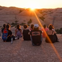 Group of people sitting in desert mountain setting at sunset, with backs facing the camera