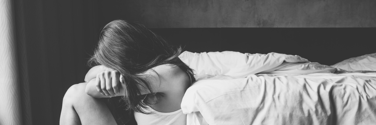 What It Feels Like to Go Nonverbal as a Verbal Autistic An upset nonverbal woman sits on the floor by her bed, her knees are pulled up and her arms are wresting on them, and her face is hidden in arms.