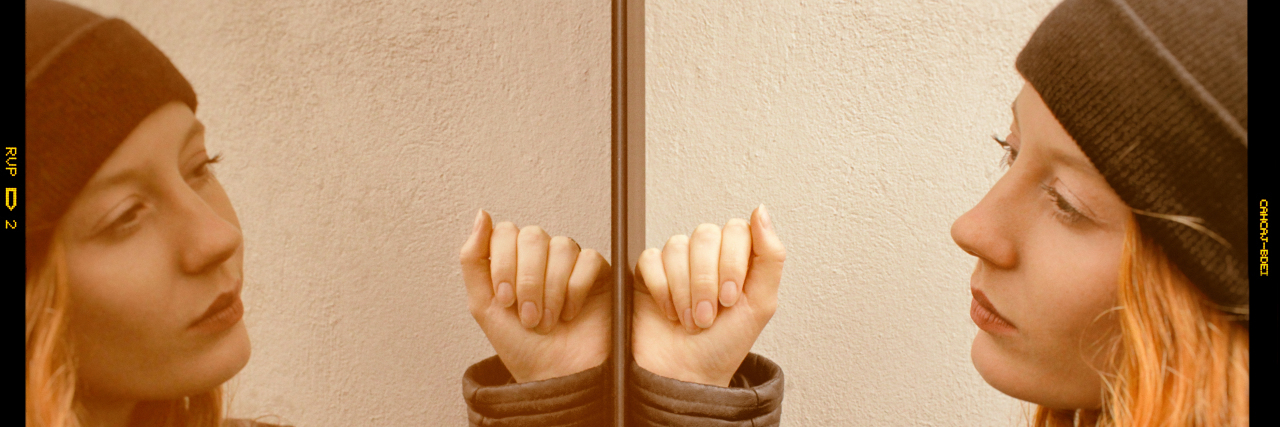 Processing Trauma and Memories on Your Surgery Anniversary Woman with long red hair wearing a black woolen cap and a black leather jacket looking at herself mirrored on a glass door with a thoughtful expression.