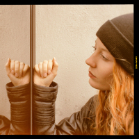 Woman with long red hair wearing a black woolen cap and a black leather jacket looking at herself mirrored on a glass door with a thoughtful expression.