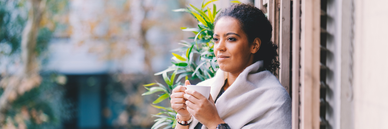 Grounding Exercises to Cope With Thanksgiving Mental Health Triggers Woman of color, looking peaceful and standing on terrace wearing a wrap and holding cup of coffee