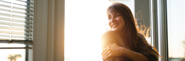 Woman standing next to window, smiling with sunlight coming in
