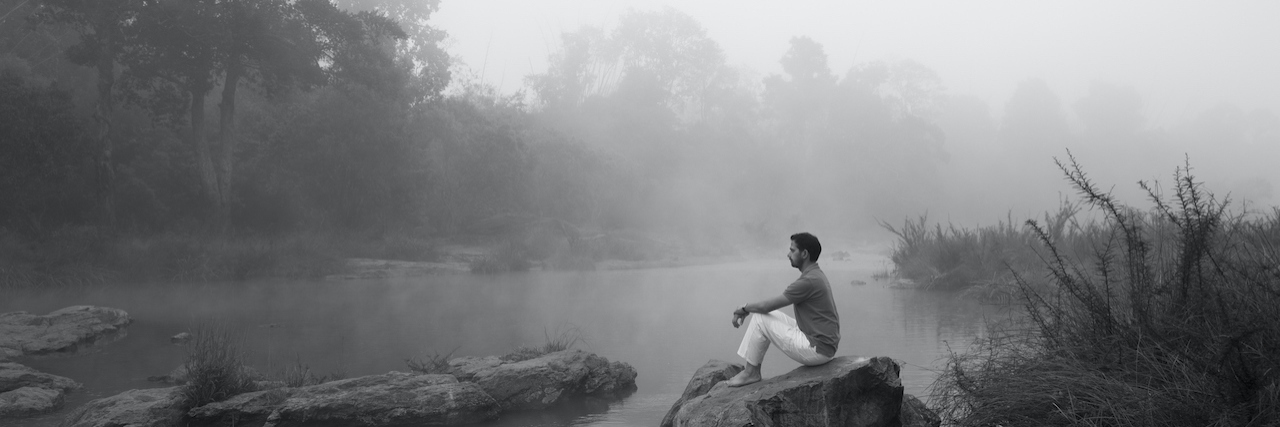 What People With Lyme Disease Need to Know Black and white photo of man staring in front of him while sitting on a rock surrounded by water and fog