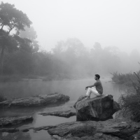 Black and white photo of man staring in front of him while sitting on a rock surrounded by water and fog