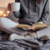Woman holding cup of tea and reading in bed. Decorative lights in background.