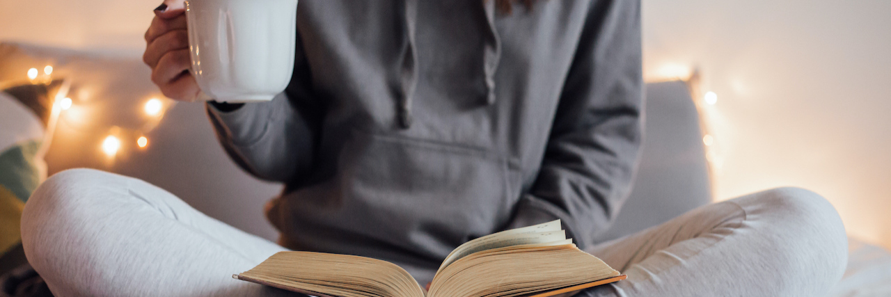How I'm Protecting My Mental Health Against Holiday Exhaustion Woman holding cup of tea and reading in bed. Decorative lights in background.