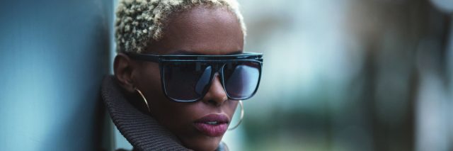 Black woman with short blonde hair in sunglasses and a black leather jacket, looking over her shoulder