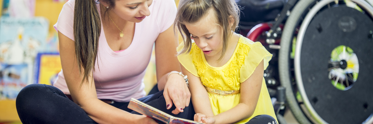 8 Awesome Children's Books About Disability A young girl is sitting in front of her wheelchair. She is in an elementary school classroom with her helper. They are reading a story book together on the floor..