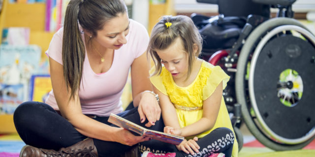 8 Awesome Children's Books About Disability A young girl is sitting in front of her wheelchair. She is in an elementary school classroom with her helper. They are reading a story book together on the floor..