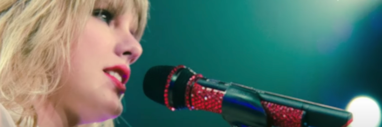 Taylor Swift Re-Claiming Her Music is Poignant For Trauma Survivors Screen grab from Taylor Swift's lyric video "Red" (Taylor's Version). She's against a blue backdrop with a red microphone in hand.