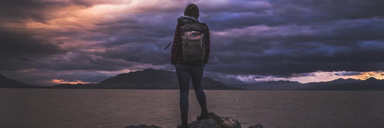 I Struggled to Practice Gratitude Until I Accepted My Grief photo of a person standing on rock in front of water and dramatic stormy skies