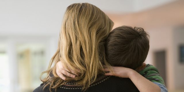 Photo from behind of a blonde woman in a black sweater holding a white, brunette young boy whose arms are wrapped around her and whose head is rested on her shoulder.