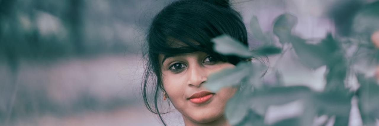 Woman of color peeking out behind plant with a slight smile