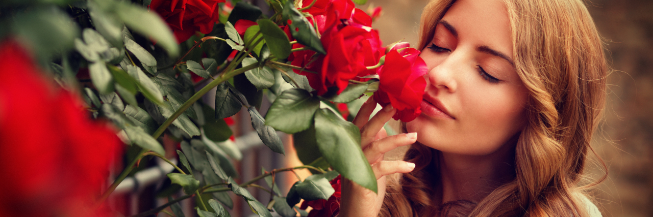 Learning to 'Smell the Roses' in Life With Chronic Pain A woman with curly reddish-blonde hair faces a rose bush with her eyes closed and smells a red rose.