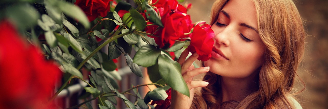 A woman with curly reddish-blonde hair faces a rose bush with her eyes closed and smells a red rose.